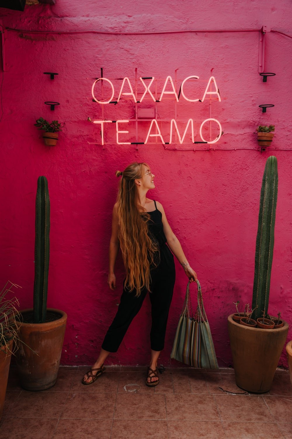 Blonde female digital nomad posing for photo in front of pink wall with a sign that reads, "Oaxaca Te Amo" in Oaxaca City, Mexico.